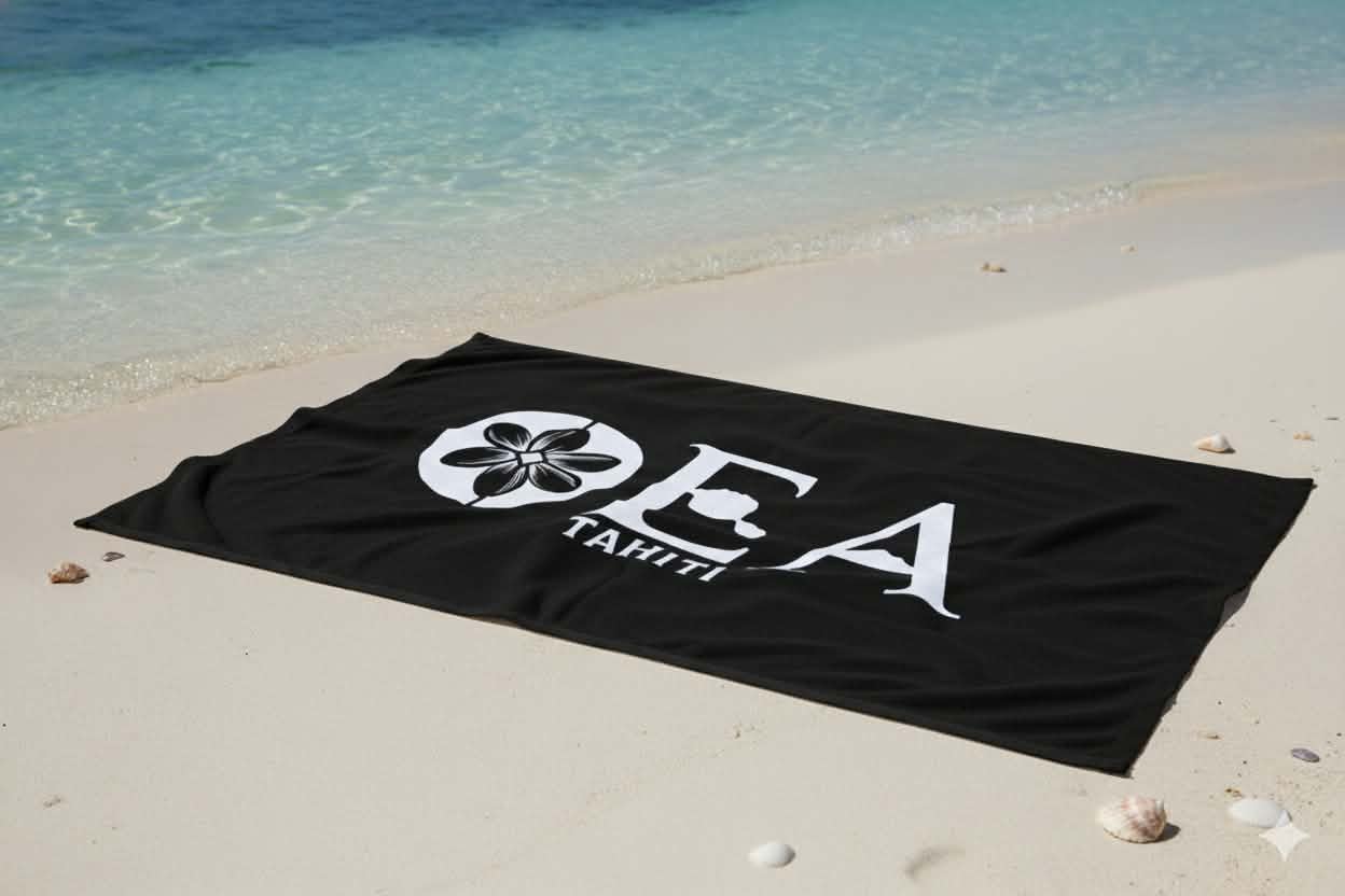 Serviette de plage OEA noire, logo complet, posée sur le sable face au lagon
